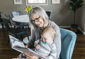 woman with gray hair wearing reading glasses and reading to infant