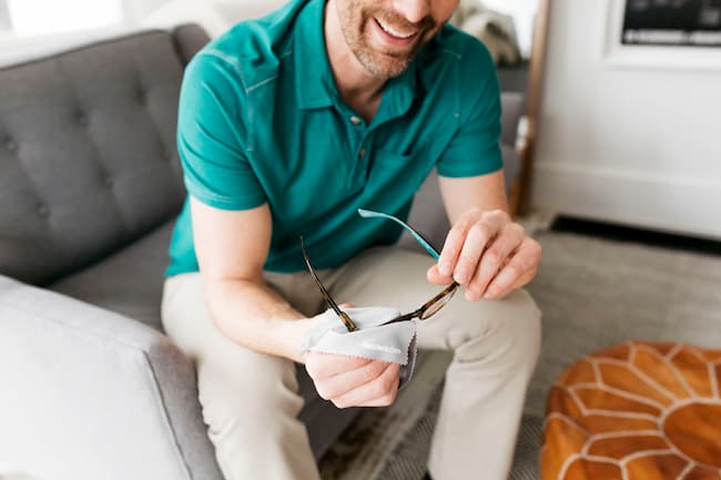 smiling man on couch cleaning glasses with cloth